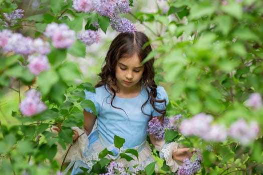 Young caucasian girl in blue dress surrounded by blooming lilacs in garden.