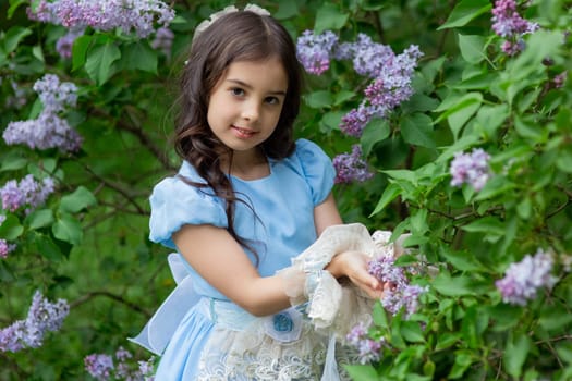 Young caucasian girl in blue dress amidst blooming lilacs.