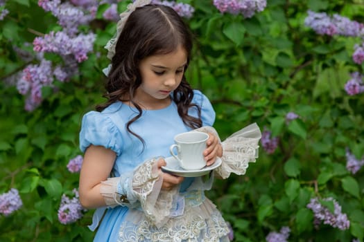 Caucasian child in blue dress enjoying tea in garden with lilac flowers.