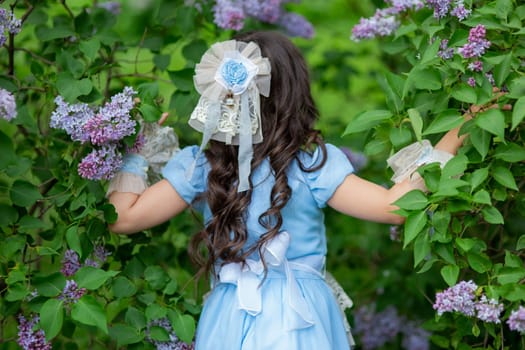 Young caucasian girl in blue dress with long hair among lilac bushes.