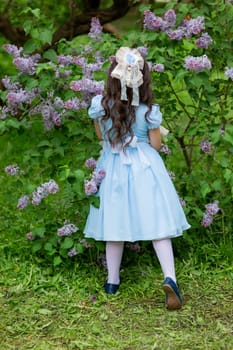 Young girl in blue dress with white bow standing by lilac bush in garden.