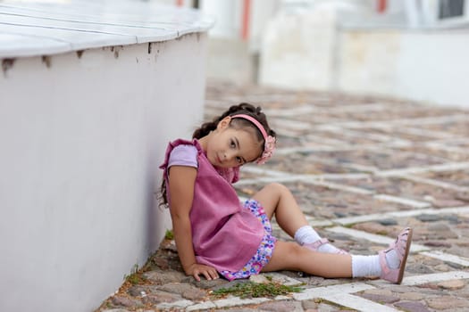 Young pretty girl in pink dress sitting on cobblestone path outdoors.