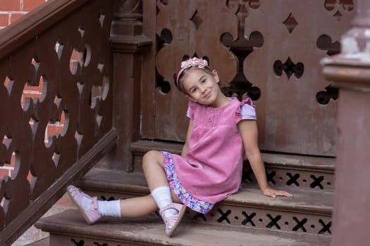 Cute young girl in pink dress sitting on wooden stairs smiling.