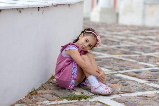 Young hispanic girl sitting on cobblestone street wearing pink dress and headband.