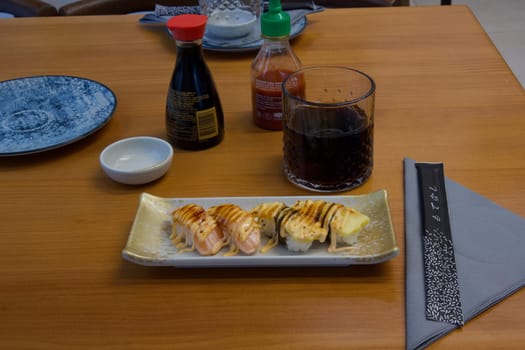 A plate of sushi sits on a wooden table with a napkin folded into a triangle. The table is set with condiments and a glass of wine. Scene is relaxed and inviting