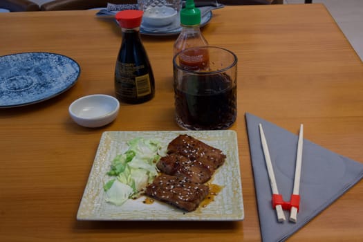 A plate of meat and vegetables sits on a wooden table with chopsticks and condiments. The table setting is simple and elegant, with a focus on the food
