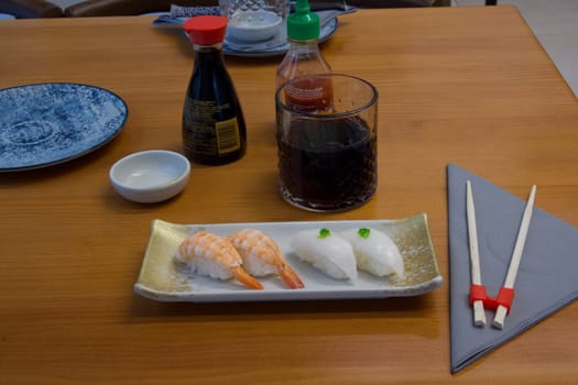 A plate of sushi with a glass of soda and chopsticks on a table. Scene is casual and relaxed, as it is a typical setting for enjoying a meal with friends or family