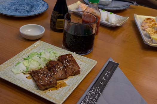 A plate of meat and vegetables is placed on a wooden table with a napkin and a cup of soda. The table is surrounded by other plates of food and condiments, creating a cozy