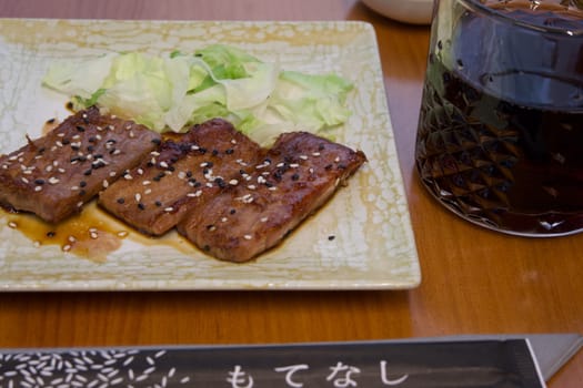 A plate of meat with sesame seeds and a glass of dark liquid. The plate is on a wooden table