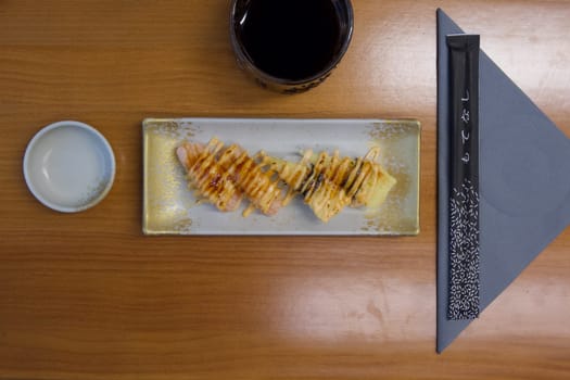 A plate of sushi with sauce and a cup of coffee on a wooden table. The tablecloth is a blue and white striped cloth