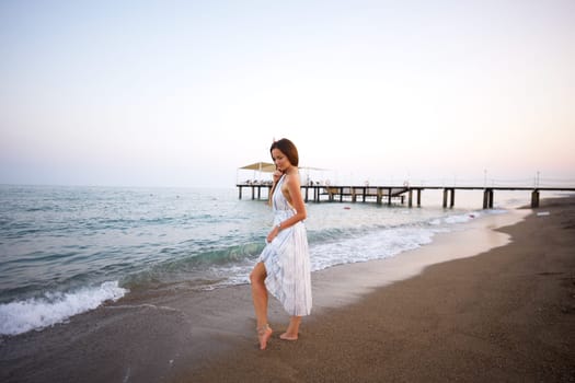 A beautiful woman wearing a light, flowing summer dress stands barefoot on a tranquil sandy beach at dusk, near gentle waves and a wooden pier.