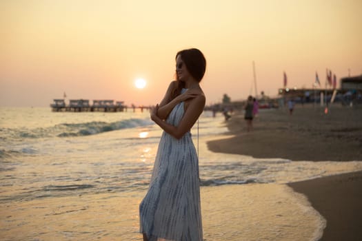 A peaceful sunset view of a woman walking barefoot on a sandy beach, surrounded by gentle waves, glowing skies, and a tranquil pier, creating a dreamy and relaxing seaside atmosphere.