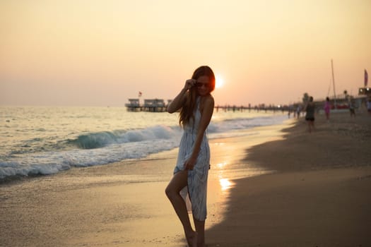 A serene beach scene featuring a woman in a flowing dress walking at sunset, with calm waves, soft sand, and a picturesque pier bathed in warm golden light, evoking peace and natural beauty