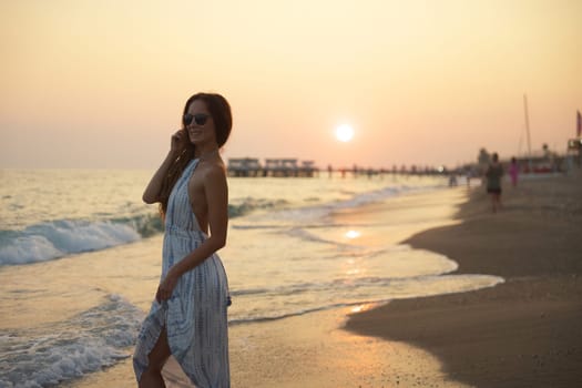 A tranquil beach scene of a woman smiling as she walks along the shoreline at sunset, with flowing dress, gentle waves, and a pier in the background enhancing the peaceful seaside ambiance.