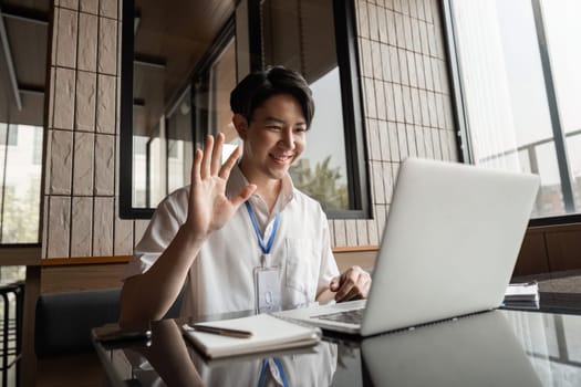 A young professional smiles and waves during a video call while using a laptop in a stylish office environment, showcasing engagement and communication.