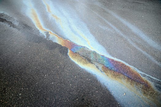 An overhead view of an asphalt road with a spreading, colorful gasoline slick creating a significant polluted area