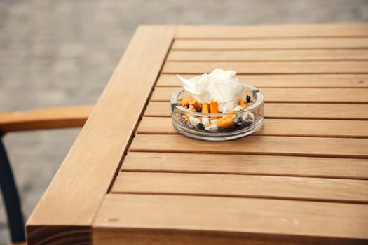 A glass ashtray filled with cigarette butts and crumpled tissues sits on a wooden outdoor table. The image highlights smoking culture, pollution, and public space hygiene