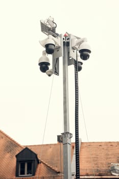 A surveillance pole with multiple security cameras and a warning system, standing in front of a historic European building. A representation of modern security in urban spaces