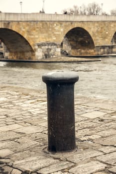 A worn mooring bollard on a historic cobblestone riverbank, set against the backdrop of an ancient stone bridge. Ideal for themes of history, travel, and urban heritage
