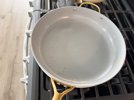 Castle Rock, Colorado, USA-January 17, 2024-A white ceramic pan with gold handles sits on a gas stove in a contemporary kitchen The stove features a stainless steel finish with multiple burners and control knobs A backsplash with white hexagonal tiles is visible in the background along with a utensil holder containing various kitchen tools and a bottle of cooking oil The scene showcases a clean and stylish cooking space