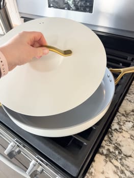 Castle Rock, Colorado, USA-January 17, 2024-A white ceramic pan with gold handles sits on a gas stove in a contemporary kitchen The stove features a stainless steel finish with multiple burners and control knobs A backsplash with white hexagonal tiles is visible in the background along with a utensil holder containing various kitchen tools and a bottle of cooking oil The scene showcases a clean and stylish cooking space