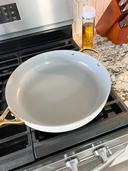 Castle Rock, Colorado, USA-January 17, 2024-A white ceramic pan with gold handles sits on a gas stove in a contemporary kitchen The stove features a stainless steel finish with multiple burners and control knobs A backsplash with white hexagonal tiles is visible in the background along with a utensil holder containing various kitchen tools and a bottle of cooking oil The scene showcases a clean and stylish cooking space
