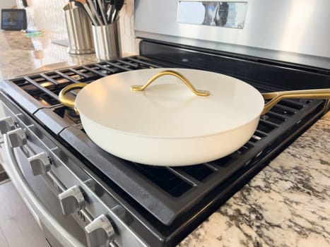 Castle Rock, Colorado, USA-January 17, 2024-A white ceramic pan with gold handles sits on a gas stove in a contemporary kitchen The stove features a stainless steel finish with multiple burners and control knobs A backsplash with white hexagonal tiles is visible in the background along with a utensil holder containing various kitchen tools and a bottle of cooking oil The scene showcases a clean and stylish cooking space