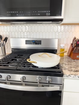 Castle Rock, Colorado, USA-January 17, 2024-A white ceramic pan with gold handles sits on a gas stove in a contemporary kitchen The stove features a stainless steel finish with multiple burners and control knobs A backsplash with white hexagonal tiles is visible in the background along with a utensil holder containing various kitchen tools and a bottle of cooking oil The scene showcases a clean and stylish cooking space