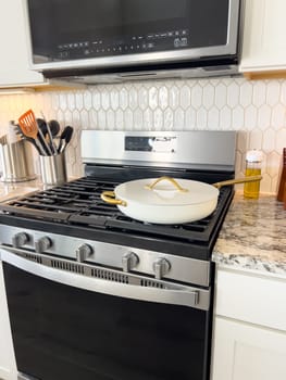 Castle Rock, Colorado, USA-January 17, 2024-A white ceramic pan with gold handles sits on a gas stove in a contemporary kitchen The stove features a stainless steel finish with multiple burners and control knobs A backsplash with white hexagonal tiles is visible in the background along with a utensil holder containing various kitchen tools and a bottle of cooking oil The scene showcases a clean and stylish cooking space