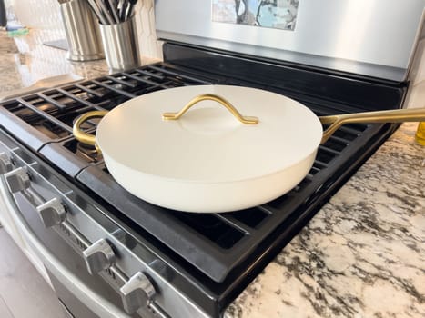 Castle Rock, Colorado, USA-January 17, 2024-A white ceramic pan with gold handles sits on a gas stove in a contemporary kitchen The stove features a stainless steel finish with multiple burners and control knobs A backsplash with white hexagonal tiles is visible in the background along with a utensil holder containing various kitchen tools and a bottle of cooking oil The scene showcases a clean and stylish cooking space