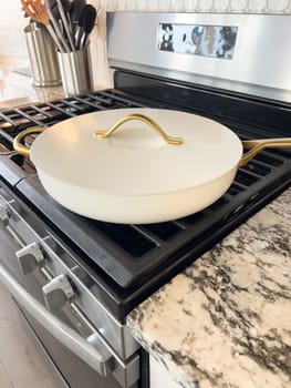 Castle Rock, Colorado, USA-January 17, 2024-A white ceramic pan with gold handles sits on a gas stove in a contemporary kitchen The stove features a stainless steel finish with multiple burners and control knobs A backsplash with white hexagonal tiles is visible in the background along with a utensil holder containing various kitchen tools and a bottle of cooking oil The scene showcases a clean and stylish cooking space