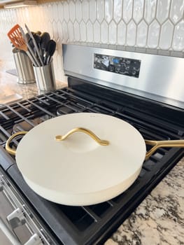 Castle Rock, Colorado, USA-January 17, 2024-A white ceramic pan with gold handles sits on a gas stove in a contemporary kitchen The stove features a stainless steel finish with multiple burners and control knobs A backsplash with white hexagonal tiles is visible in the background along with a utensil holder containing various kitchen tools and a bottle of cooking oil The scene showcases a clean and stylish cooking space
