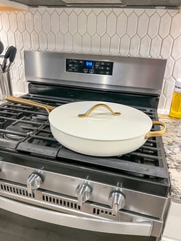 Castle Rock, Colorado, USA-January 17, 2024-A white ceramic pan with gold handles sits on a gas stove in a contemporary kitchen The stove features a stainless steel finish with multiple burners and control knobs A backsplash with white hexagonal tiles is visible in the background along with a utensil holder containing various kitchen tools and a bottle of cooking oil The scene showcases a clean and stylish cooking space