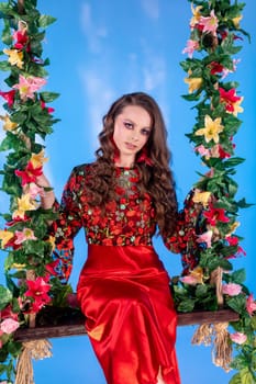 A young woman in a colorful outfit sits gracefully on a floral swing against a bright blue backdrop