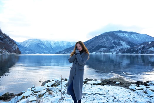 Young woman standing by a serene snow-covered lake in Gaupne, Norway, surrounded by mountains, enjoying the peaceful winter landscape