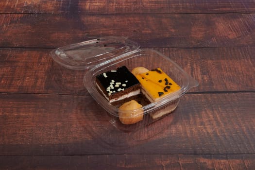 A box of assorted desserts sits on a wooden table. The desserts include a chocolate cake, a donut, and a piece of fruit. The box is open, revealing the delicious treats inside