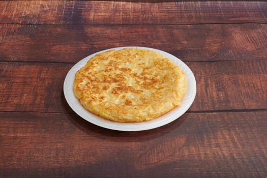 A plate of food with a yellowish brown crust sits on a wooden table. The plate is white and the food is round