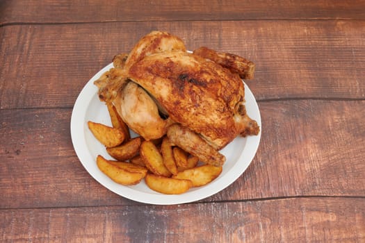 A plate of chicken and potatoes on a wooden table. The chicken is cooked and has a crispy skin. The potatoes are cut into wedges and are seasoned with salt and pepper