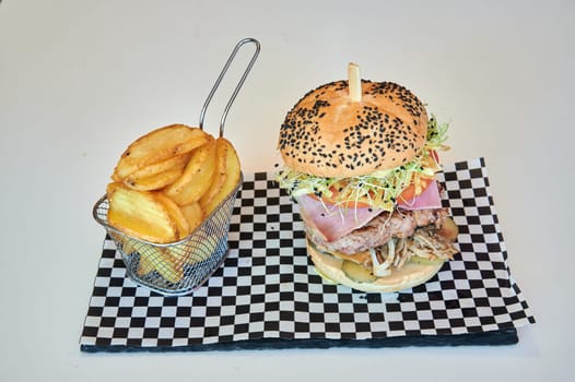 A burger with lettuce and tomato sits on a black and white checkered paper plate next to a basket of french fries