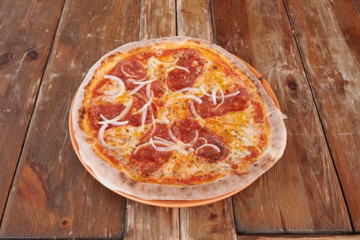 A pizza with pepperoni and onions on top sits on a wooden table. The pizza is cut into slices and is ready to be eaten