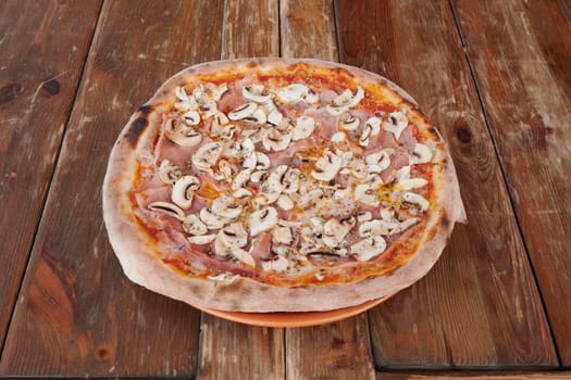 A pizza with mushrooms and ham on it sits on a wooden table. The pizza is covered in mushrooms and ham, and it looks delicious