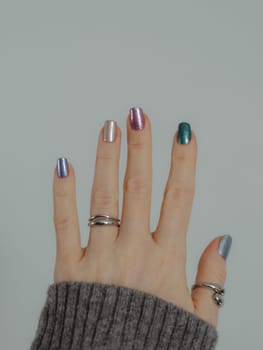 Skittles nails on hand. Colorful shiny manicure with holographic sparkle and metallic pigment. Female hand in gray sweater with multicolored nails on white wall background. Copy space. Vertical