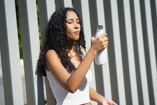 A young woman with curly hair joyfully sips from her stylish, modern water bottle, standing against a vibrant and colorful backdrop, embodying a relaxed lifestyle centered on health and joy