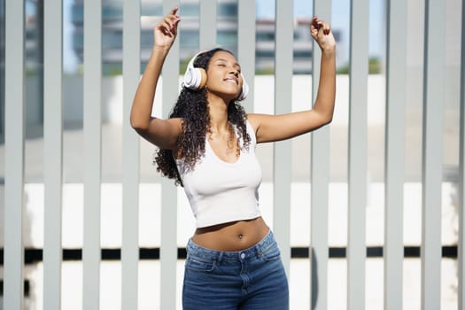 A very cheerful and lively young woman dances energetically while wearing fashionable headphones and a casual outfit, radiating pure joy and happiness against a vibrant and modern urban backdrop