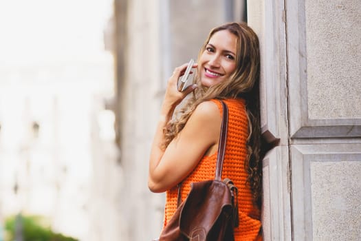 Latin woman leaning on an urban facade holding a mobile and smiling in the city