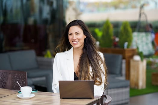 A smiling woman in a white blazer using a laptop at a caf terrace, enjoying her coffee and the outdoor ambiance