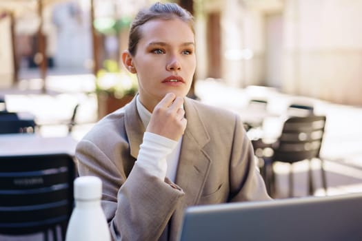 A focused woman in a beige blazer sits outdoors, contemplating while using her laptop in a caf setting