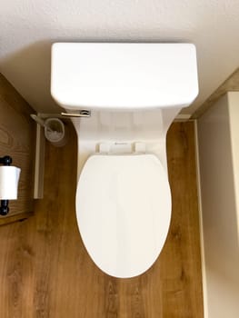 Broomfield, Colorado, USA-January 12, 2025-A clean white toilet with a closed lid is installed in a small bathroom with wood-style flooring The toilet is positioned against a white wall with a toilet paper holder mounted on the side and a small trash can nearby A bathtub is partially visible on the right side creating a compact yet functional bathroom space