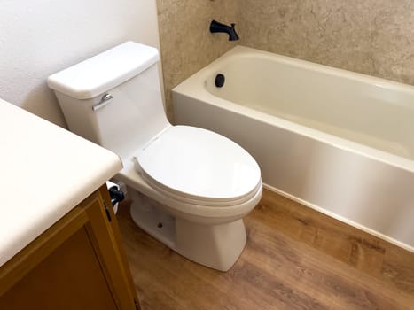 Broomfield, Colorado, USA-January 12, 2025-A clean white toilet with a closed lid is installed in a small bathroom with wood-style flooring The toilet is positioned against a white wall with a toilet paper holder mounted on the side and a small trash can nearby A bathtub is partially visible on the right side creating a compact yet functional bathroom space