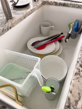 Castle Rock, Colorado, USA-January 18, 2024-A white kitchen sink filled with dirty dishes including plates bowls a coffee mug and utensils such as tongs and knives A dish rack with soapy water is placed on one side while a sponge and dish scrubber rest nearby The sink has a modern design with a garbage disposal drain